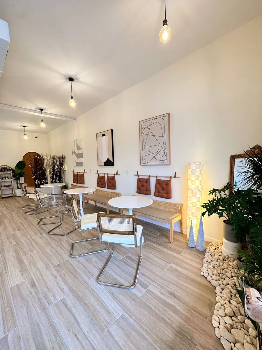 A bright, modern cafe interior with light flooring, white tables, mixed chairs, abstract art, plants, and pendant lights.