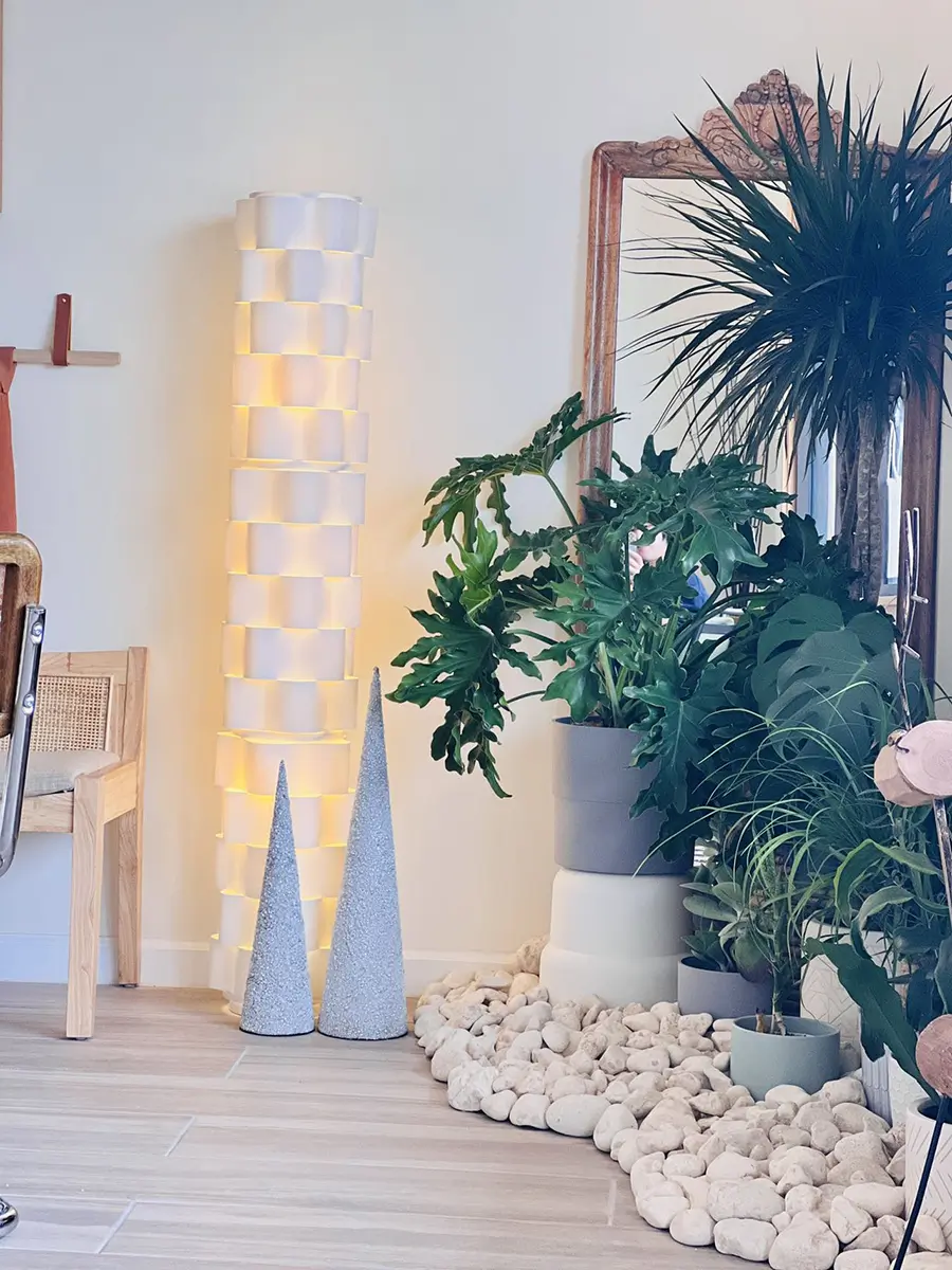 A cozy corner with a tall woven floor lamp, two cone-shaped decor pieces, various green plants in pots, a decorative mirror, and a rocky accent area on light wood flooring.